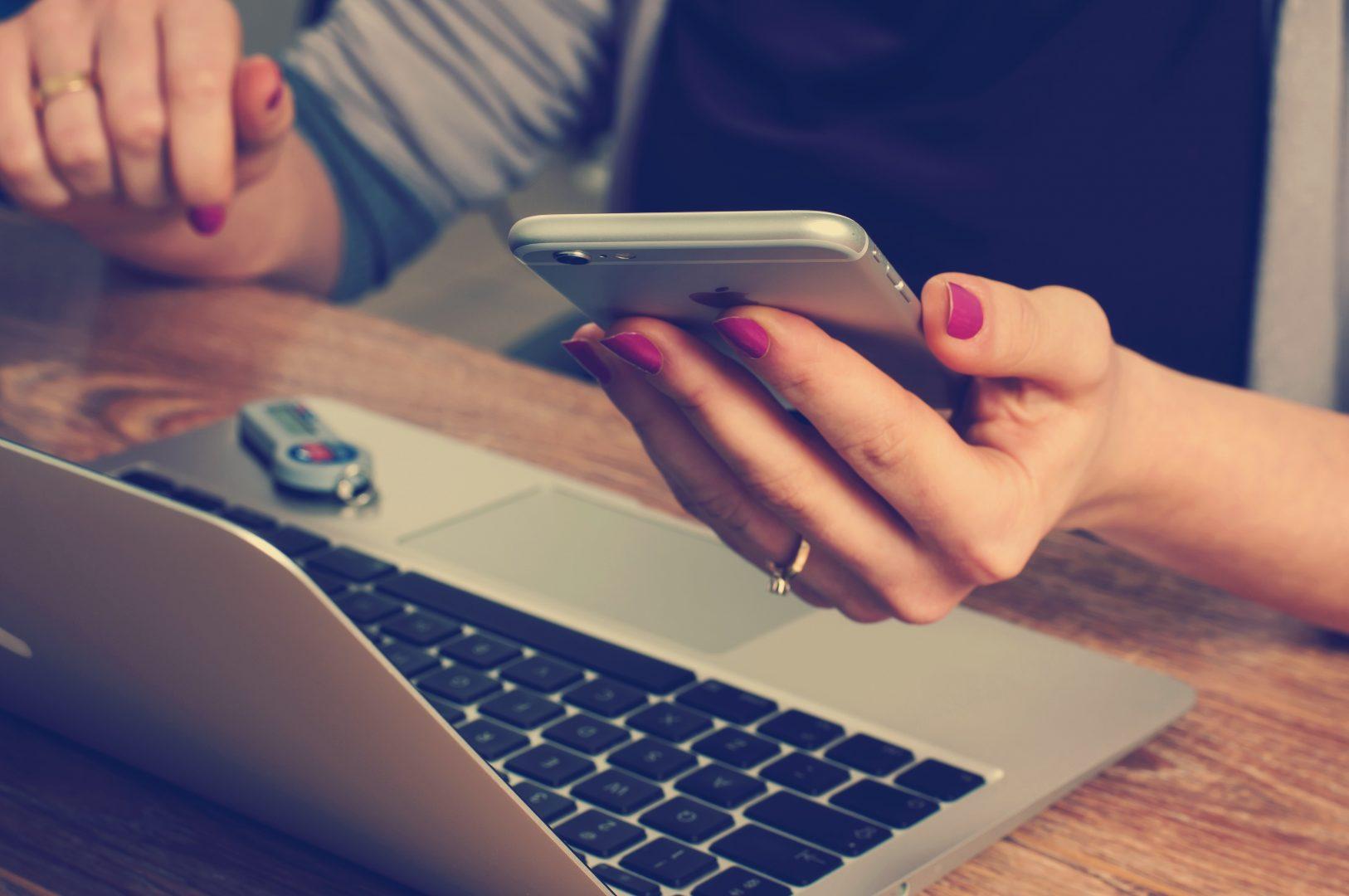 Someone holding a phone whilst using a laptop, a way to use digital marketing in Surrey.