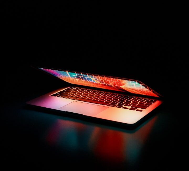 An almost closed laptop lit with bright colour against a black background