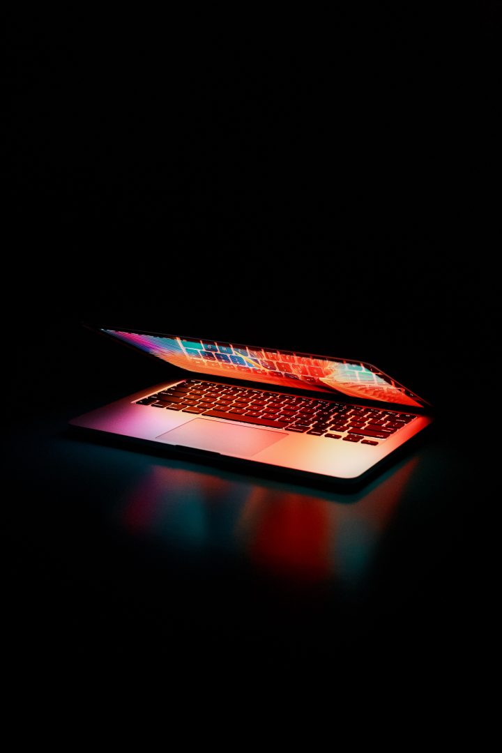 An almost closed laptop lit with bright colour against a black background