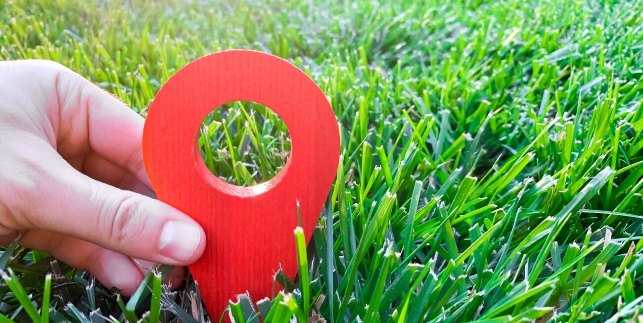 Hand holding a 3D red location pin in green grass, representing local SEO