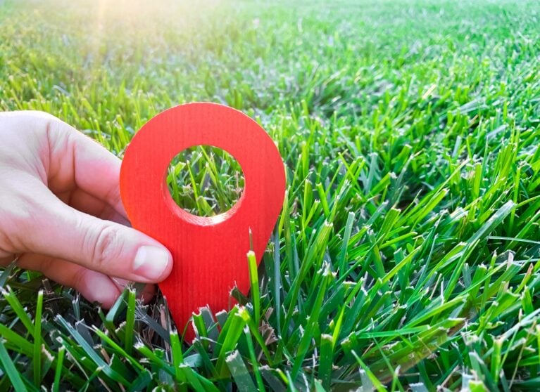 Hand holding a 3D red location pin in green grass, representing local SEO